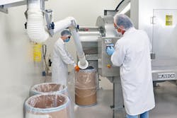 The blended batch is discharged into lined fiber drums, as dust is collected by a vacuum. A second operator adjusts the mixer to rotate slowly during discharge. The blended batch is discharged into lined fiber drums, as dust is collected by a vacuum. A second operator adjusts the mixer to rotate slowly during discharge.